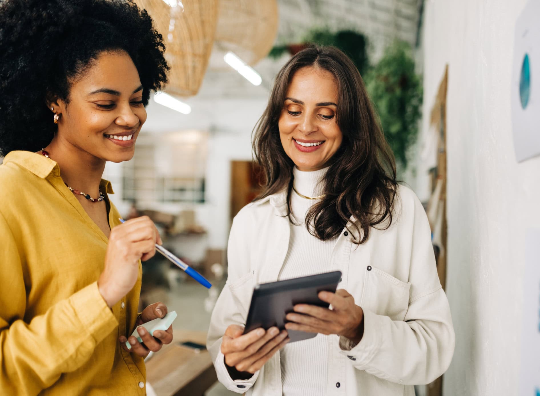 Two professional women stand and view business information on a tablet.