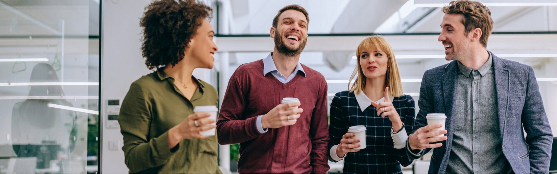 A group of 4 employees holding coffee cups walks together through a bright office