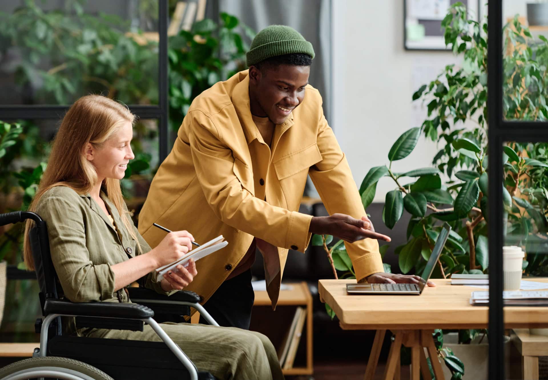 Two employees, one standing and one sitting in a wheelchair, point to a laptop to collaborate in an office filled with plants