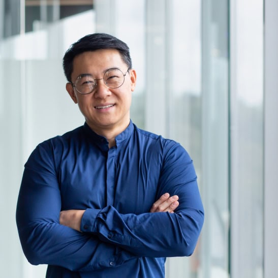A professional-looking man stands smiling with his arms folded in an office